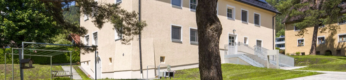 Mehrparteienhaus mit Solarmodulen auf dem Dach und einer Außentreppe, umgeben von Bäumen und Grün (Wohnanlagen, Antoniusstraße, Bludenz)