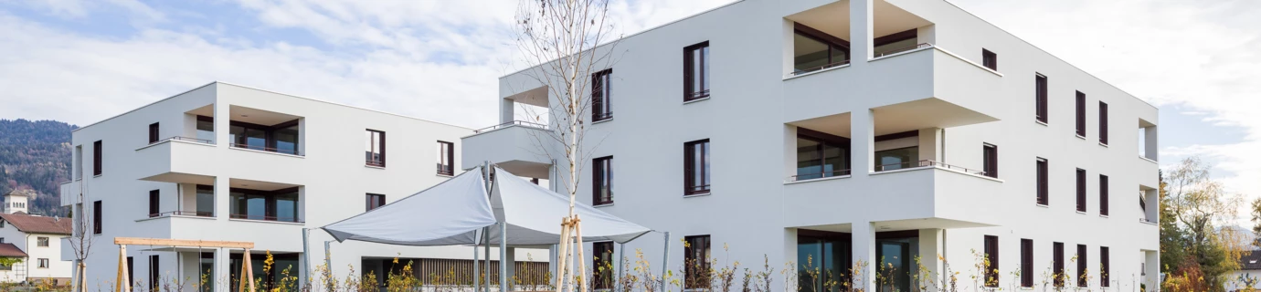 Zwei moderne weiße Wohngebäude mit Balkonen, umgeben von grünen Rasenflächen, mit einem überdachten Sitzbereich und jungen Bäumen (Unterhochstegstrasse).