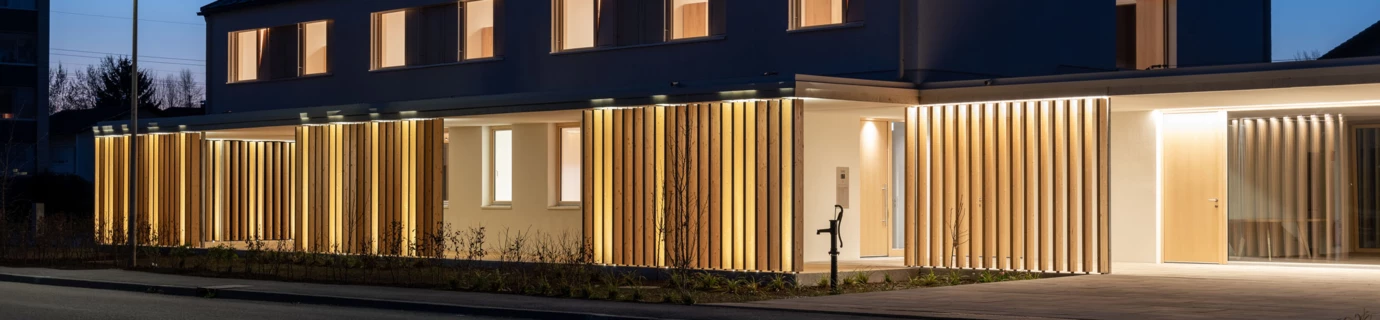 Modernes Gebäude in Wolfurt bei Nacht mit beleuchteter Holzlamellenfassade und hellen Fenstern (Dieter).