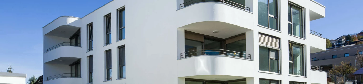 Moderne weiße Wohnanlage mit abgerundeten Balkonen, großen Fenstern, grünen Rasenflächen und jungen Bäumen unter blauem Himmel in Feldkirch (Wolf-Huber-Straße).