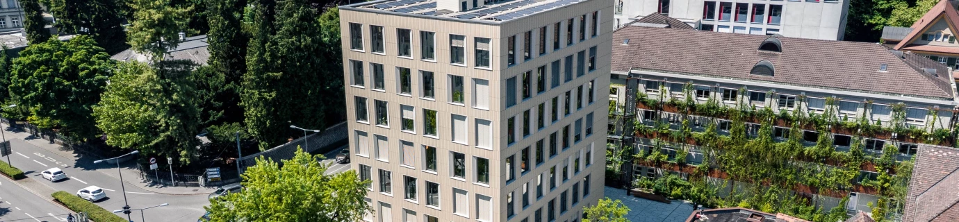 Aerial view of the modern Baerahus building with solar panels on the roof, surrounded by green trees, other buildings, and a multi-lane (Feldkirch)