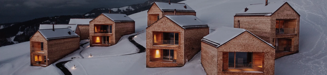 Mehrere beleuchtete Holzhäuser, sogenannte Chalets, in einer verschneiten Berglandschaft mit einem Pfad, der die Gebäude verbindet, bei Dämmerung in Furx (Bernhard).