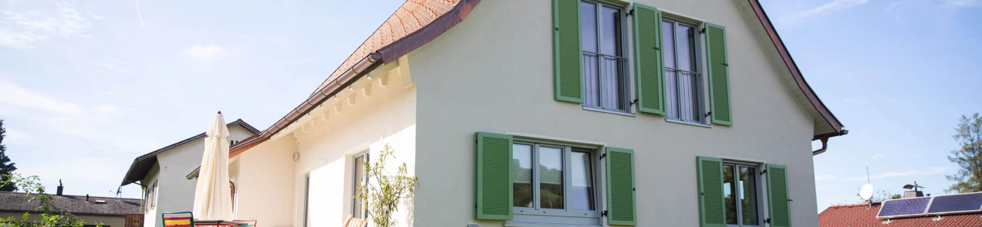 Einfamilienhaus in Dornbirn mit hellbeiger Fassade, terracottafarbenem Dach, grünen Fensterläden und einer Terrasse mit Gartenmöbeln (Generalsanierung).