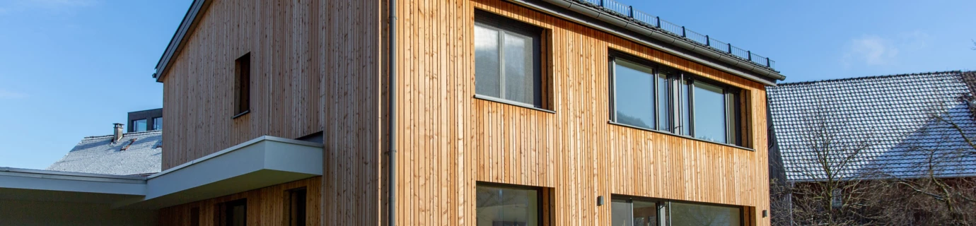 Modernes Einfamilienhaus in Dornbirn mit Holzfassade und großer Fensterfront auf einem leicht schneebedeckten Rasen unter blauem Himmel (Generalsanierung, Holzoptik).