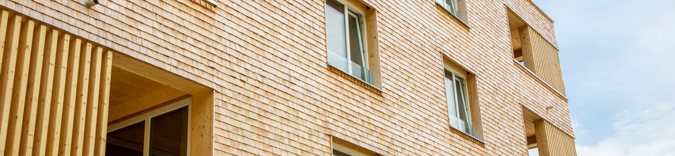 Fassadenansicht eines modernen Gebäudes mit Holzschindelverkleidung und großen Fenstern sowie hölzernen Lamellen an den Balkonen (Hellbrunnenstraße).