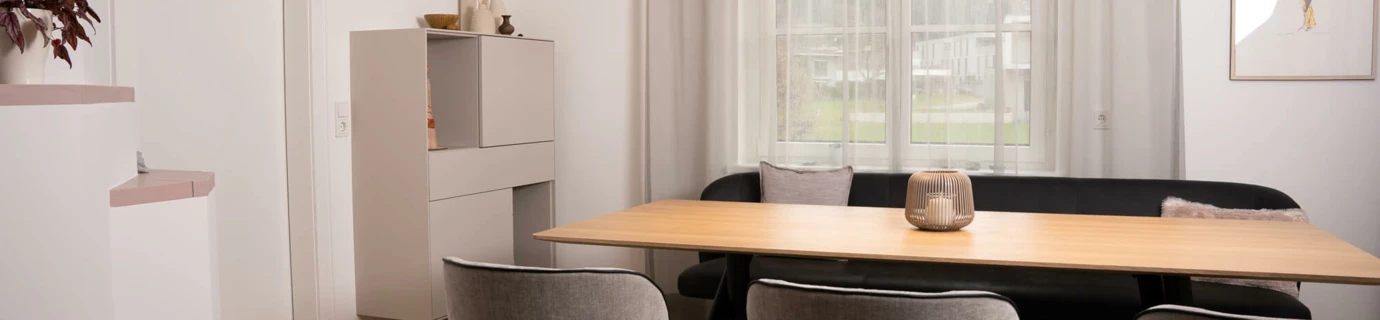 Modernes Esszimmer in einem Einfamilienhaus mit Holztisch, schwarzer Eckbank und grauen Stühlen. Helle Wandfarbe und ein Fenster lassen viel Licht herein (Generalsanierung, Bohle).