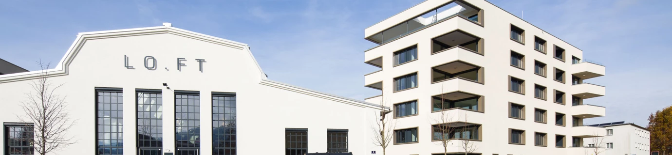 Zwei moderne Gebäude, eines mit "LO.FT" Schriftzug und großen Fenstern, sowie ein Wohnhaus mit Balkonen an einer Straße bei Tag (Bäumleareal).