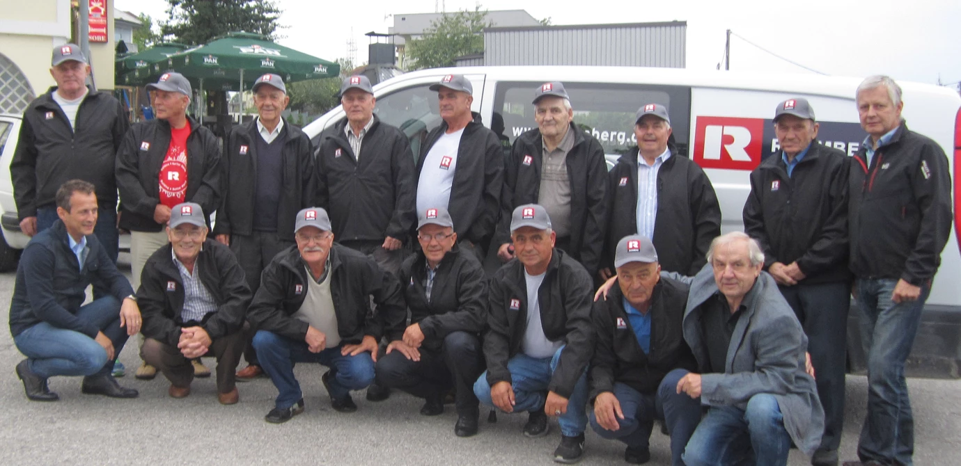A group of eighteen men of various ages, most wearing black jackets and gray Rhomberg caps, pose outdoors. Some crouch in front of a white (pensioners)