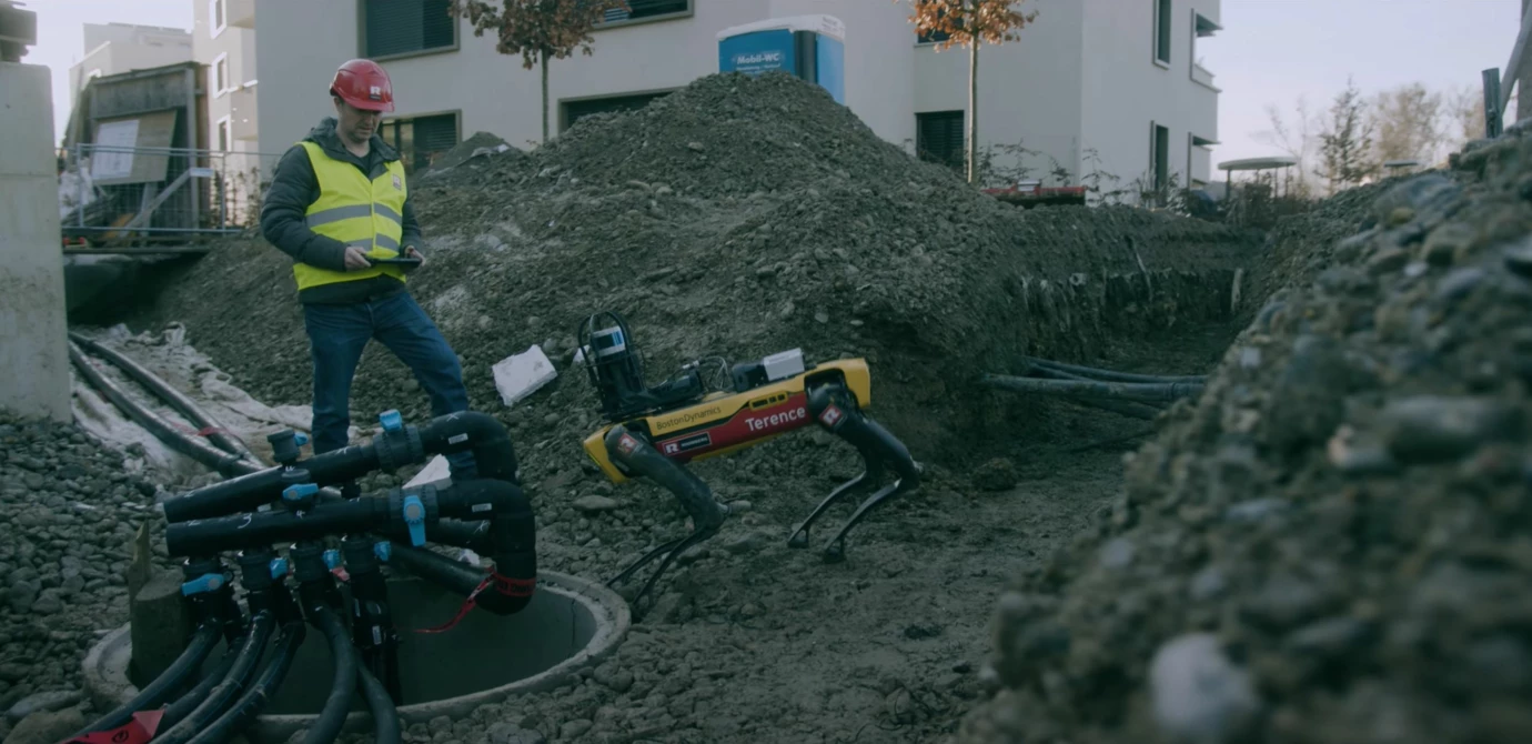 Ein Bauarbeiter in Schutzkleidung steuert einen gelben Boston Dynamics Roboterhund namens "Terence" auf einer Baustelle (Robotic, YouTube).