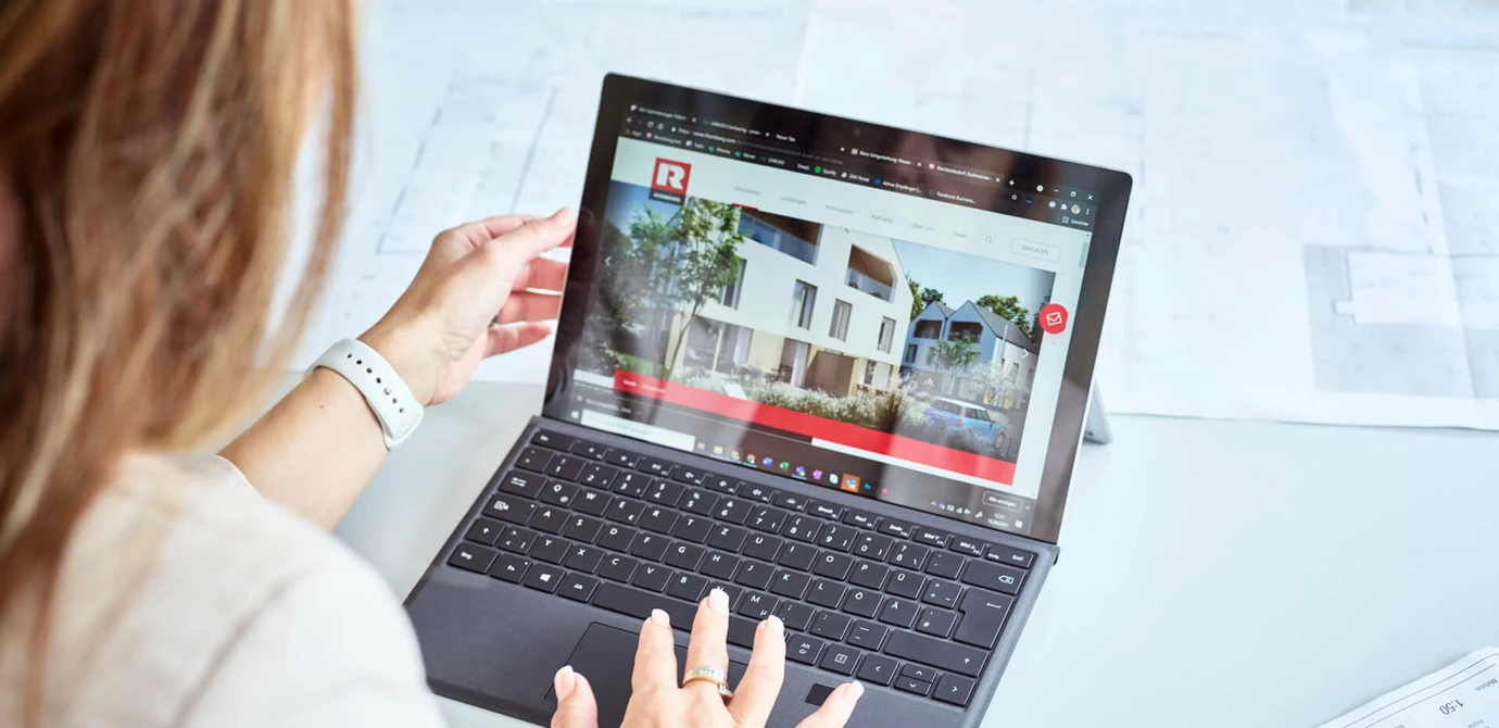 Frau arbeitet an einem Microsoft Surface Laptop mit der Rhomberg Bau Webseite, auf der ein modernes Wohngebäude abgebildet ist (Plan, Closeup, Architekt).