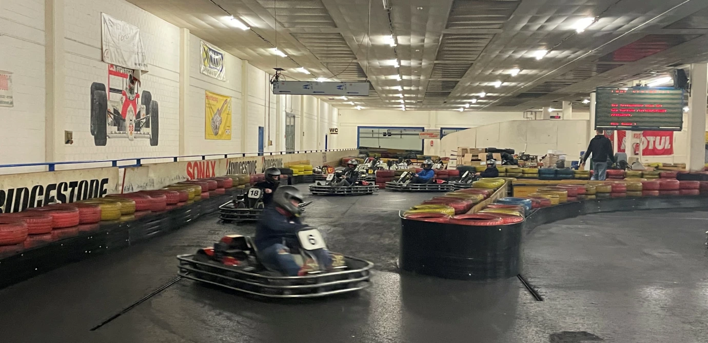 Mehrere Personen in Gokarts fahren auf einer Indoor-Rennstrecke mit Reifenbegrenzungen und Werbebannern an den Wänden (Lehrlingsabend).
