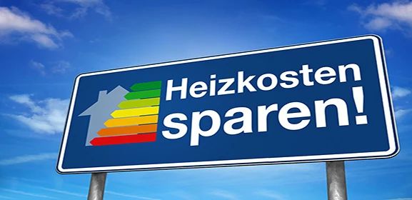 Straßenschild mit der Aufschrift "Heizkosten sparen!" und einem Haus-Symbol mit farbiger Energieskala davor, unter blauem Himmel.