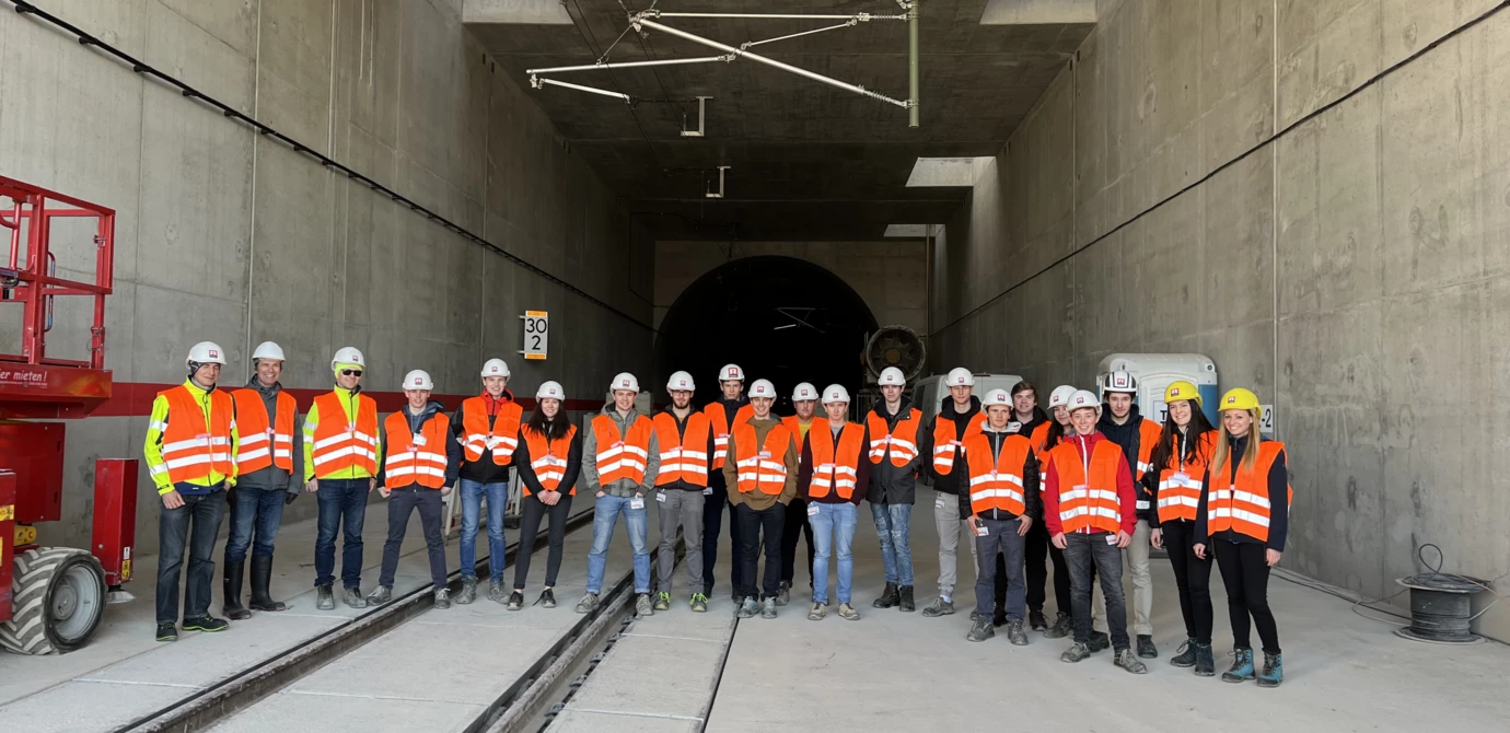 Eine Gruppe von 18 Personen in Sicherheitskleidung mit Helmen und Warnwesten steht vor einem Tunnelportal mit Gleisen (Rankweil, Deutschland, Gruppenfoto).
