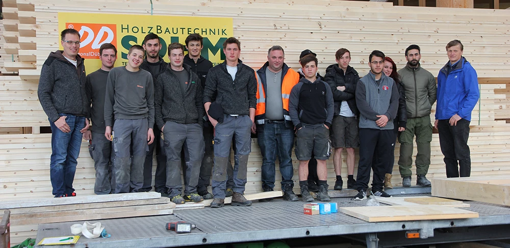 Eine Gruppe junger Männer und zwei ältere Männer posieren vor einem Stapel Holzbretter und einem Schild mit der Aufschrift "Holz Bautechnik Solm" (Lehrlingstag).