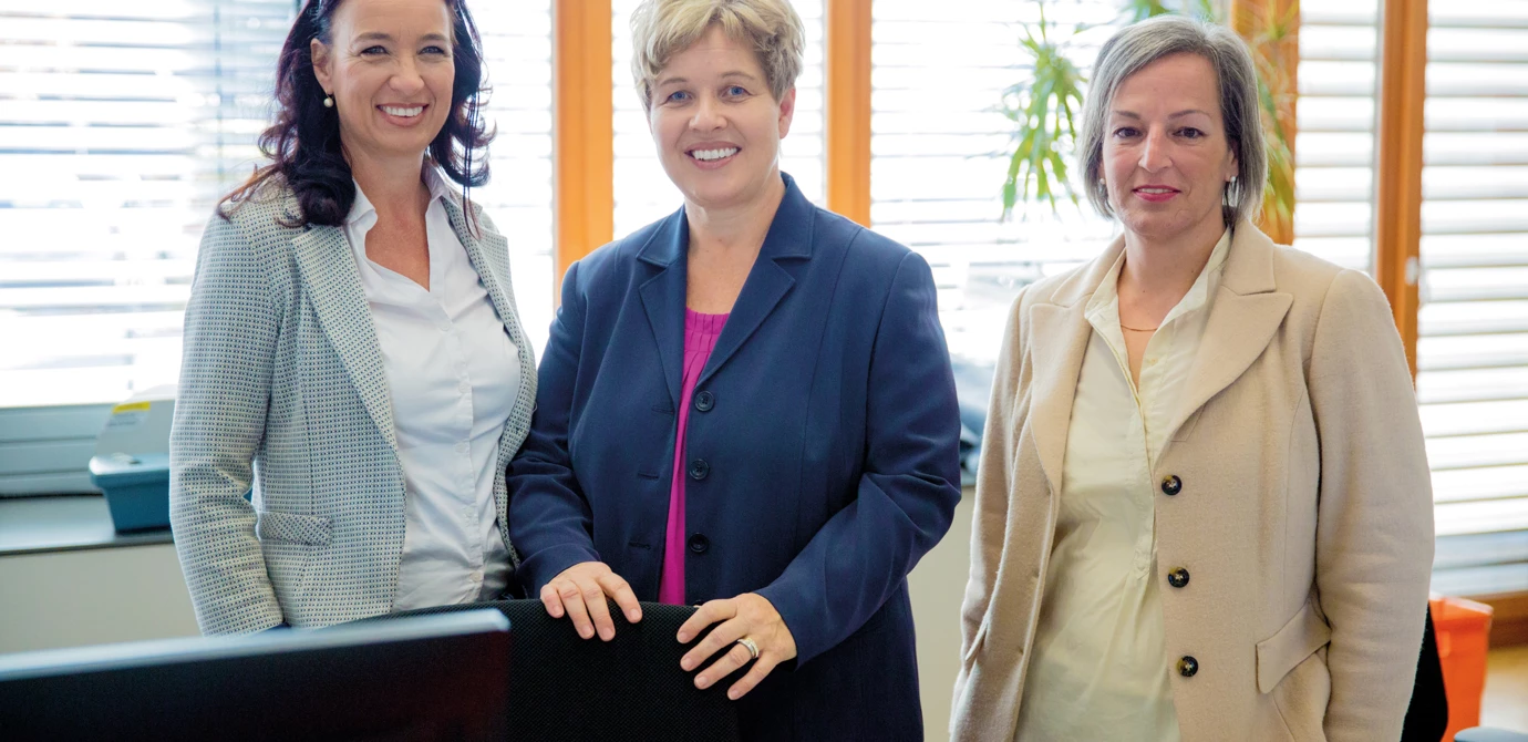 Drei lächelnde Frauen in Businesskleidung stehen vor einem Fenster in einem Büro vor einem dunklen Schreibtisch (Empfangsdamen).