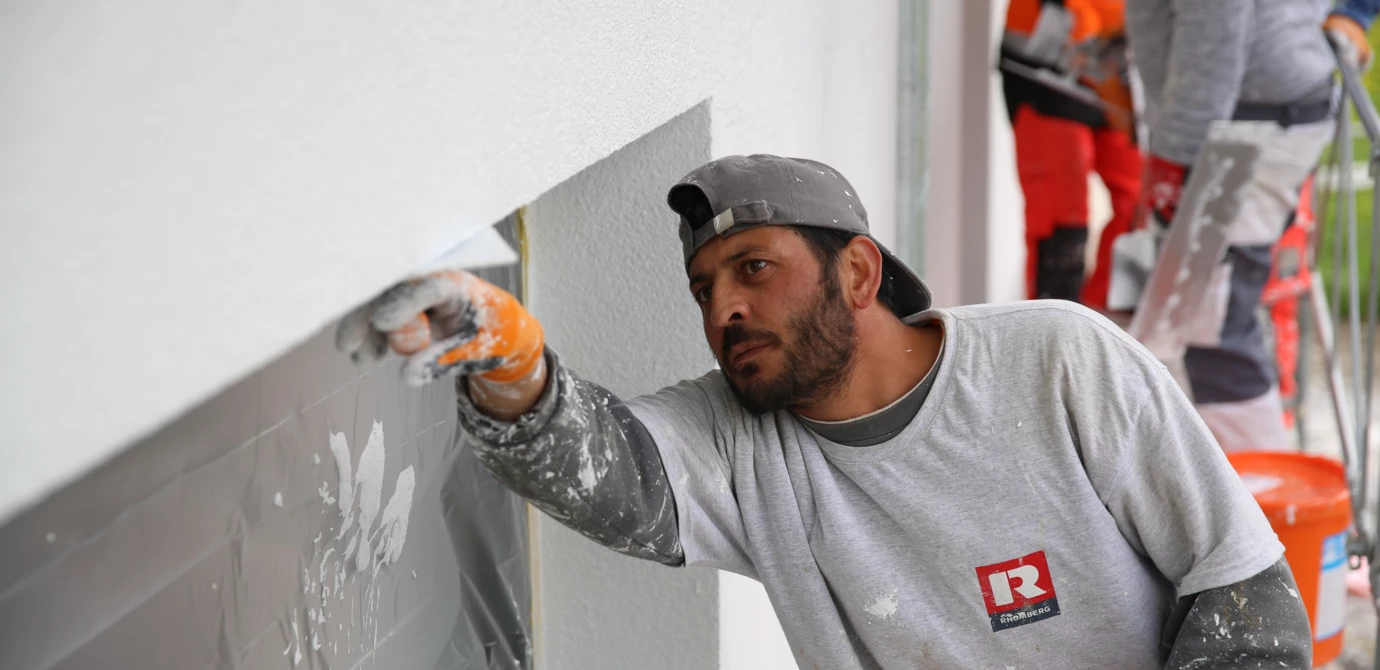 Arbeiter in Schutzkleidung verputzt eine Fensterlaibung an einem Gebäude in Meiningen, verschmutzte Bauwand (Verputzarbeiten, Hochbau)