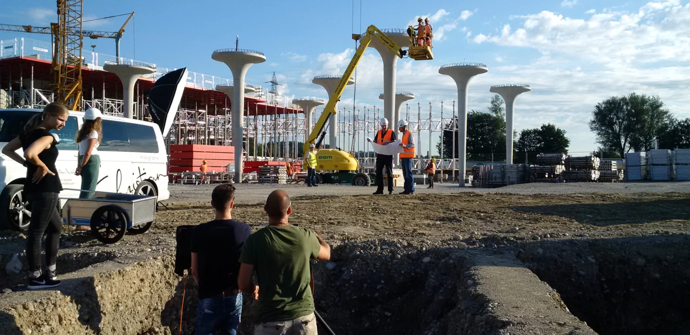 Arbeiter auf einer Baustelle mit einem Steiger, zwei Männern, die Pläne prüfen, und einer Fotografin vor einem Lieferwagen.
