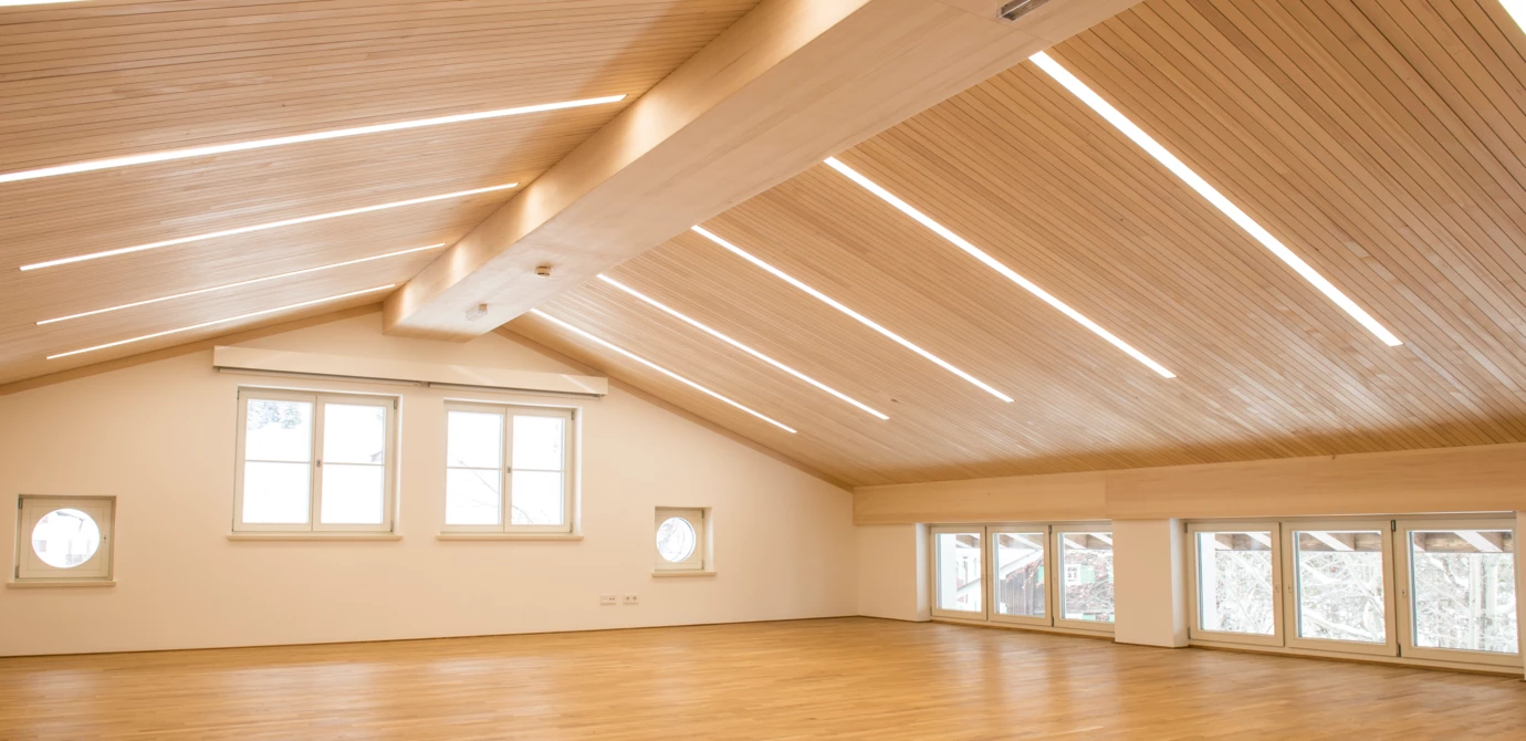 Ein großer, leerer Raum mit Holzboden und einer schrägen Holzdecke, die mit langen, linearen LED-Leuchten versehen ist (Generalsanierung, Kindes, Lech).