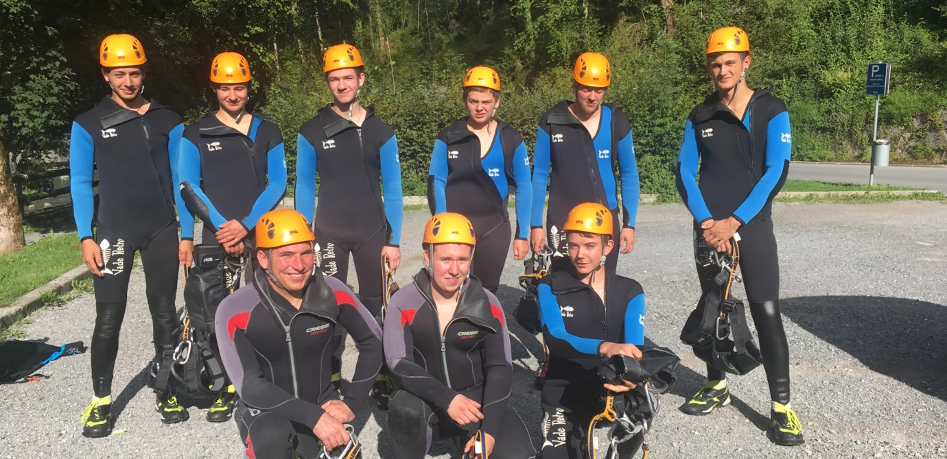 Eine Gruppe von neun jungen Erwachsenen in Neoprenanzügen und orangefarbenen Helmen, bereit für Outdoor-Aktivitäten (Lehrlingen, Rafting, Ausrüstung).