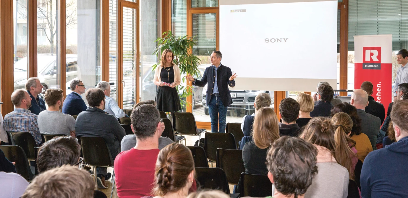 Zwei Personen präsentieren vor einem Publikum, mit Rhomberg Bau Flagge und Sony Logo an der Wand (Digital, Day).