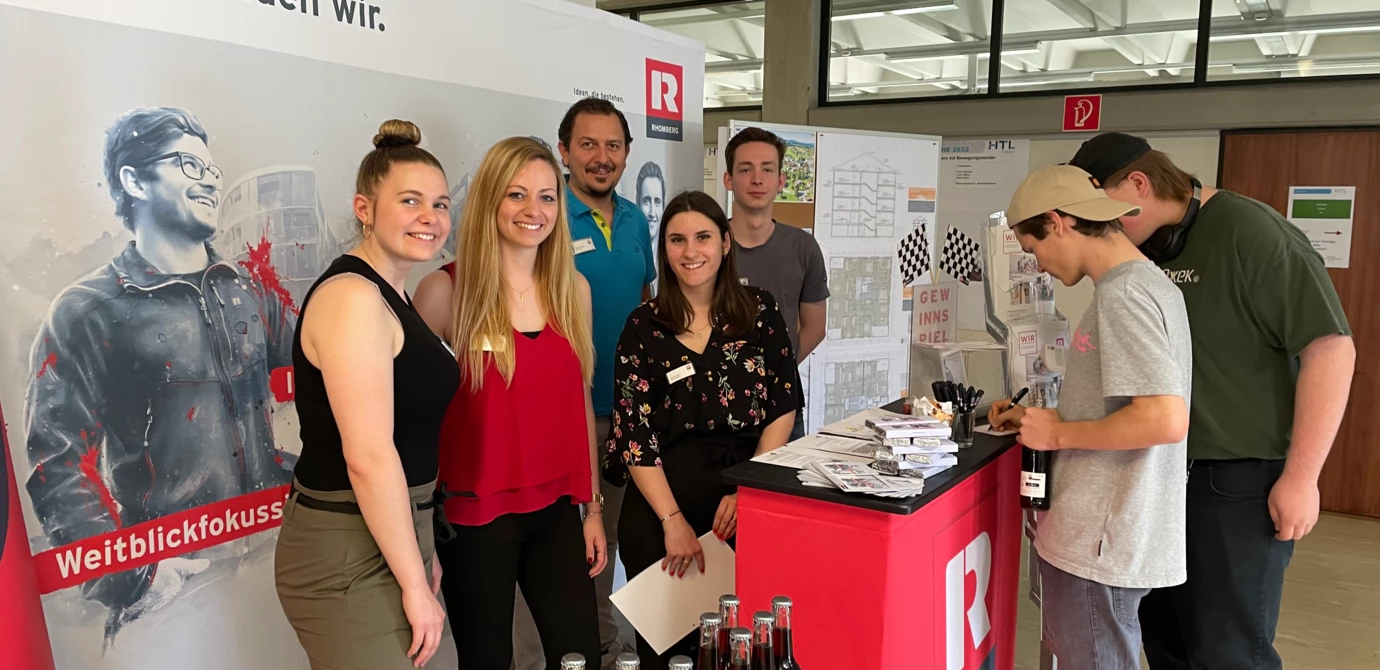 Mitarbeiter von Rhomberg Bau an einem Messestand, der "Darauf bauen wir" und "Weitblickfokus" bewirbt mit Getränken und Broschüren (Bauforum).