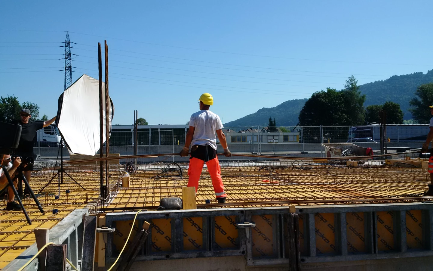 Zwei Bauarbeiter in Schutzkleidung und ein Fotograf mit Softbox auf einer Baustelle unter blauem Himmel mit Bergen und einem Hochspannungsmast im Hintergrund.