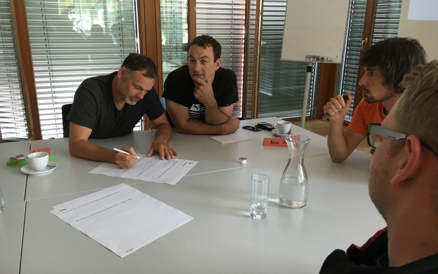 Vier Männer sitzen an einem Tisch und diskutieren bei einem Workshop, einer davon schreibt Notizen auf ein Blatt Papier.