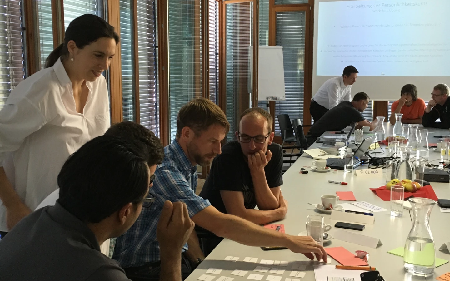 Eine Gruppe von Rhomberg Bau Mitarbeitern sitzt an einem Tisch bei einem Workshop und arrangiert Karten. Eine Präsentation ist im Hintergrund an die Wand projiziert.
