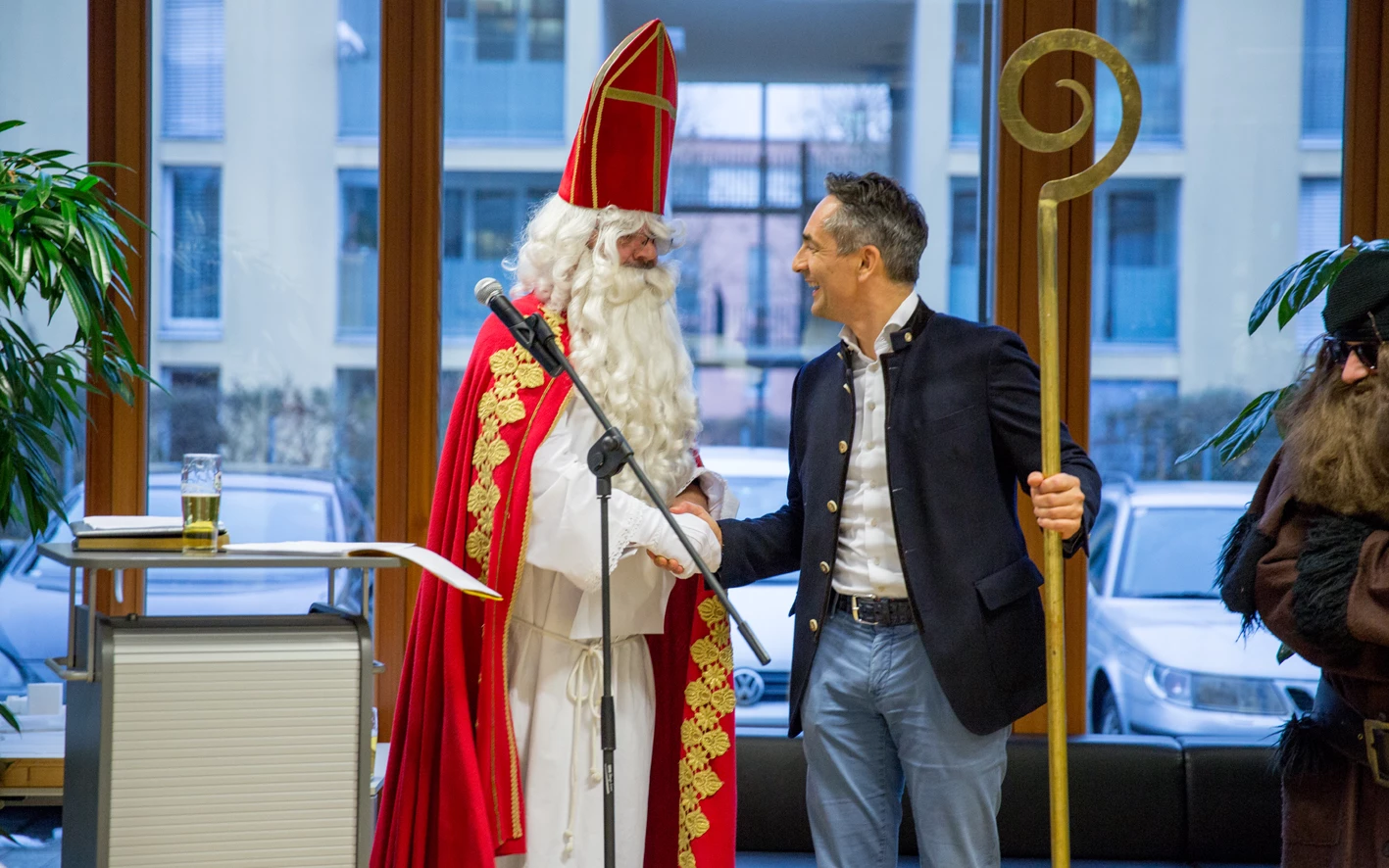 Ein lächelnder Mann im Trachtenjanker schüttelt dem Nikolaus mit Bischofsmütze, rotem Mantel und weißem Bart die Hand (Hubert, Geburtstag).