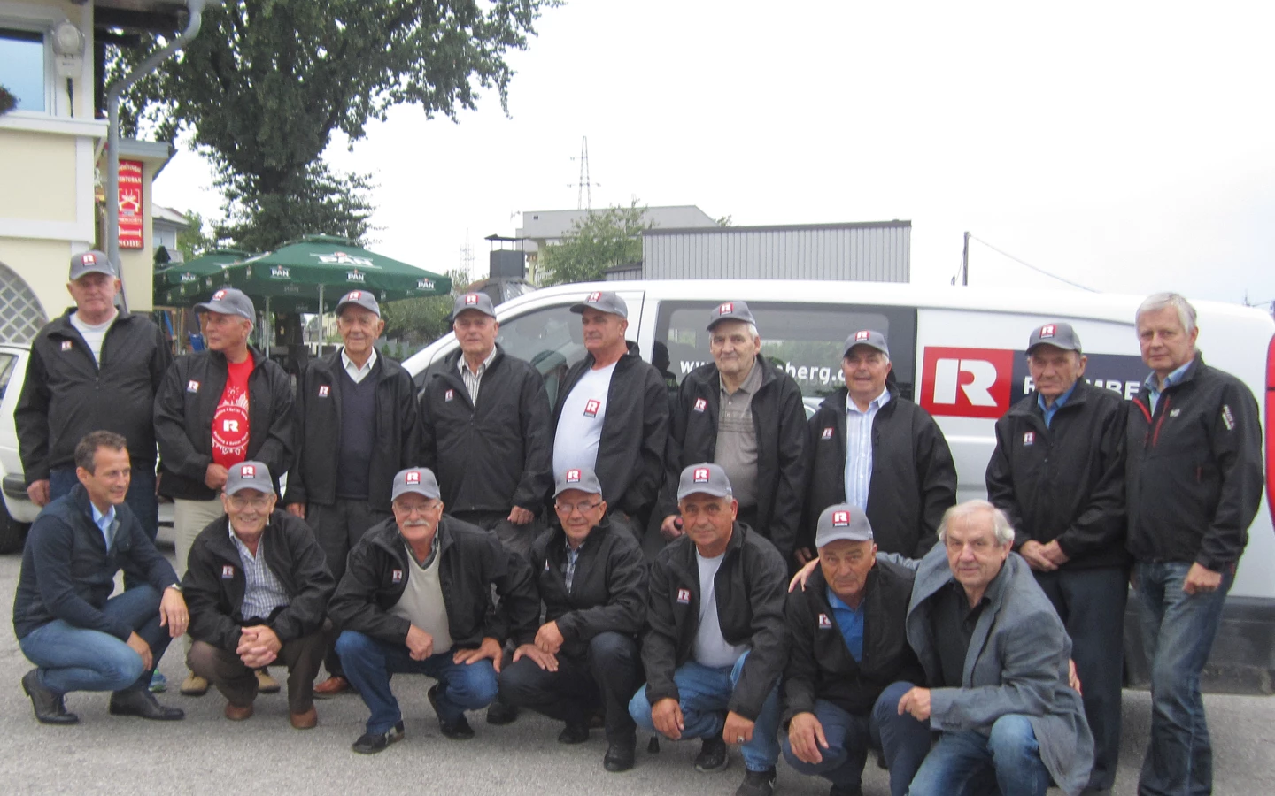 A group of eighteen men of various ages, most wearing black jackets and gray Rhomberg caps, pose outdoors. Some crouch in front of a white (pensioners)