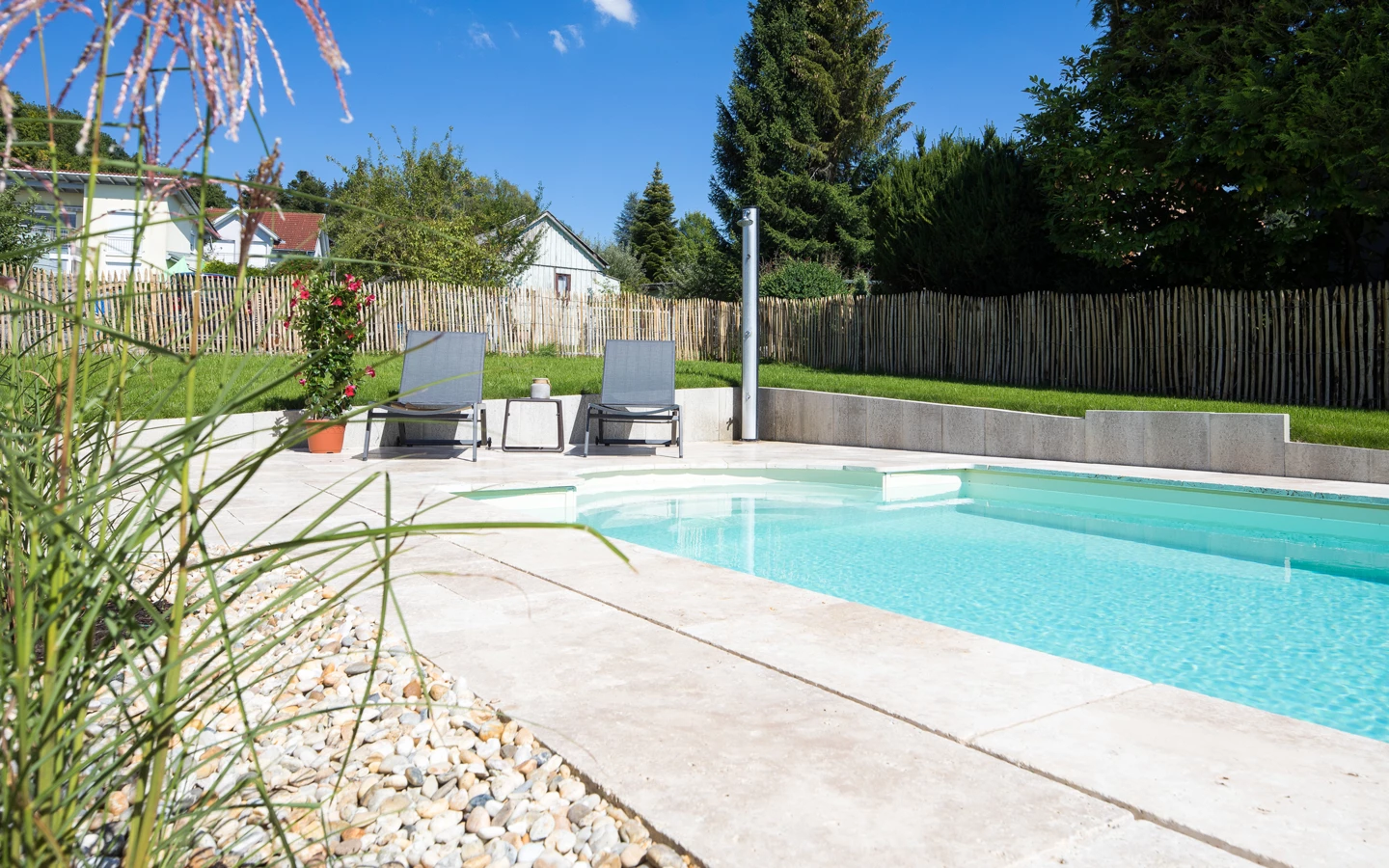 Ein rechteckiger Pool mit hellblauem Wasser, umgeben von beigem Steinbelag und zwei grauen Liegestühlen, an einem sonnigen Tag (Hohenweiler).