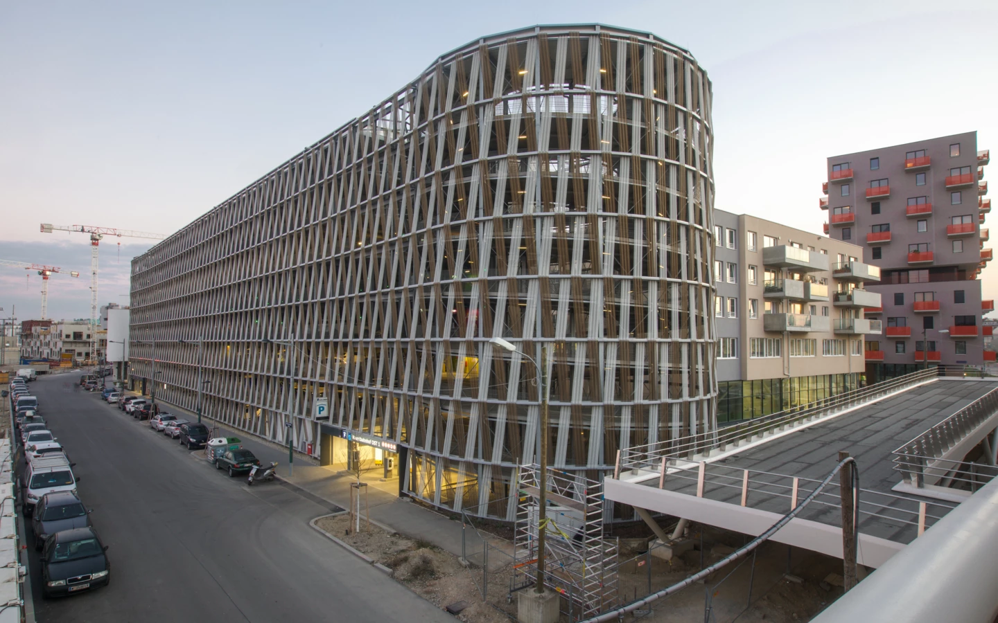 Rundes Parkhaus Wien Hauptbahnhof mit Holzlamellenfassade bei Dämmerung, umgeben von Wohngebäuden und einer Straße mit parkenden Autos (Hochgarage).