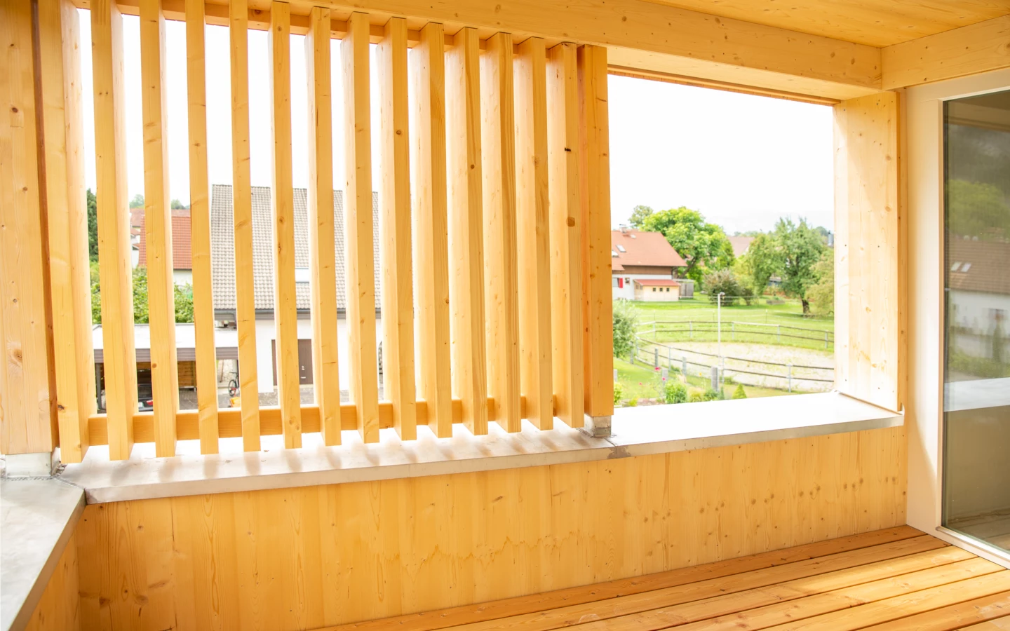 Blick von einem hölzernen Balkon mit vertikalen Lamellen und Holzfußboden auf Wohnhäuser und grüne Wiesen (Hellbrunnenstraße)