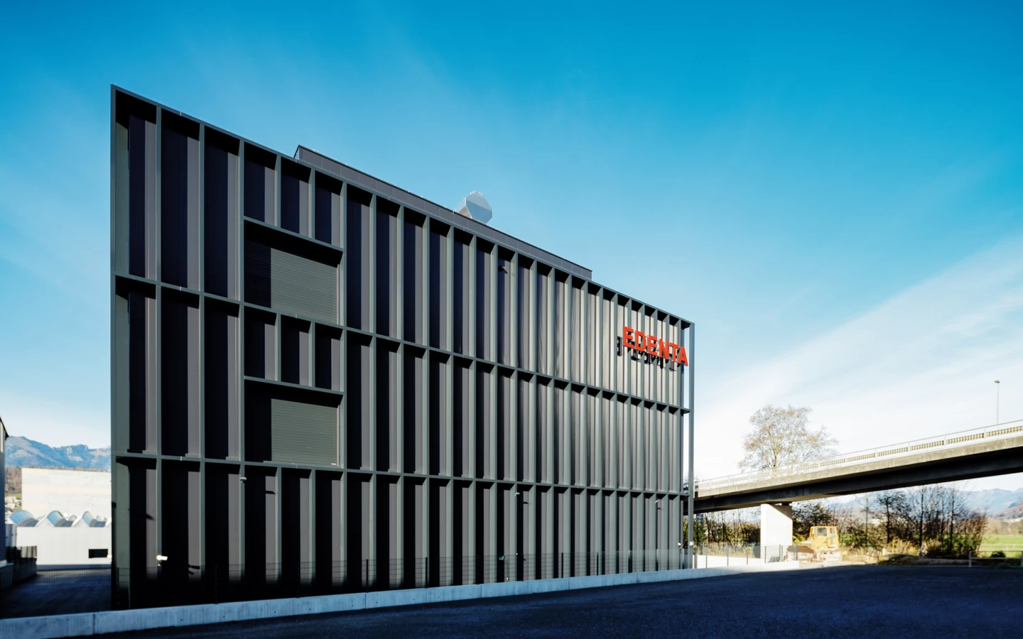 Modernes rechteckiges graues Gebäude mit rot leuchtendem EDENTA Logo an der Fassade unter blauem Himmel, mit Straßenbrücke im Hintergrund.