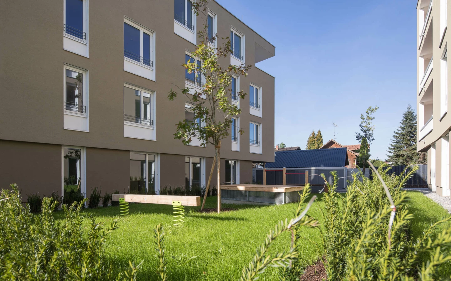 Moderne Wohnanlage Widum Lustenau mit brauner Fassade, vielen Fenstern und einem Spielplatz auf grüner Wiese.