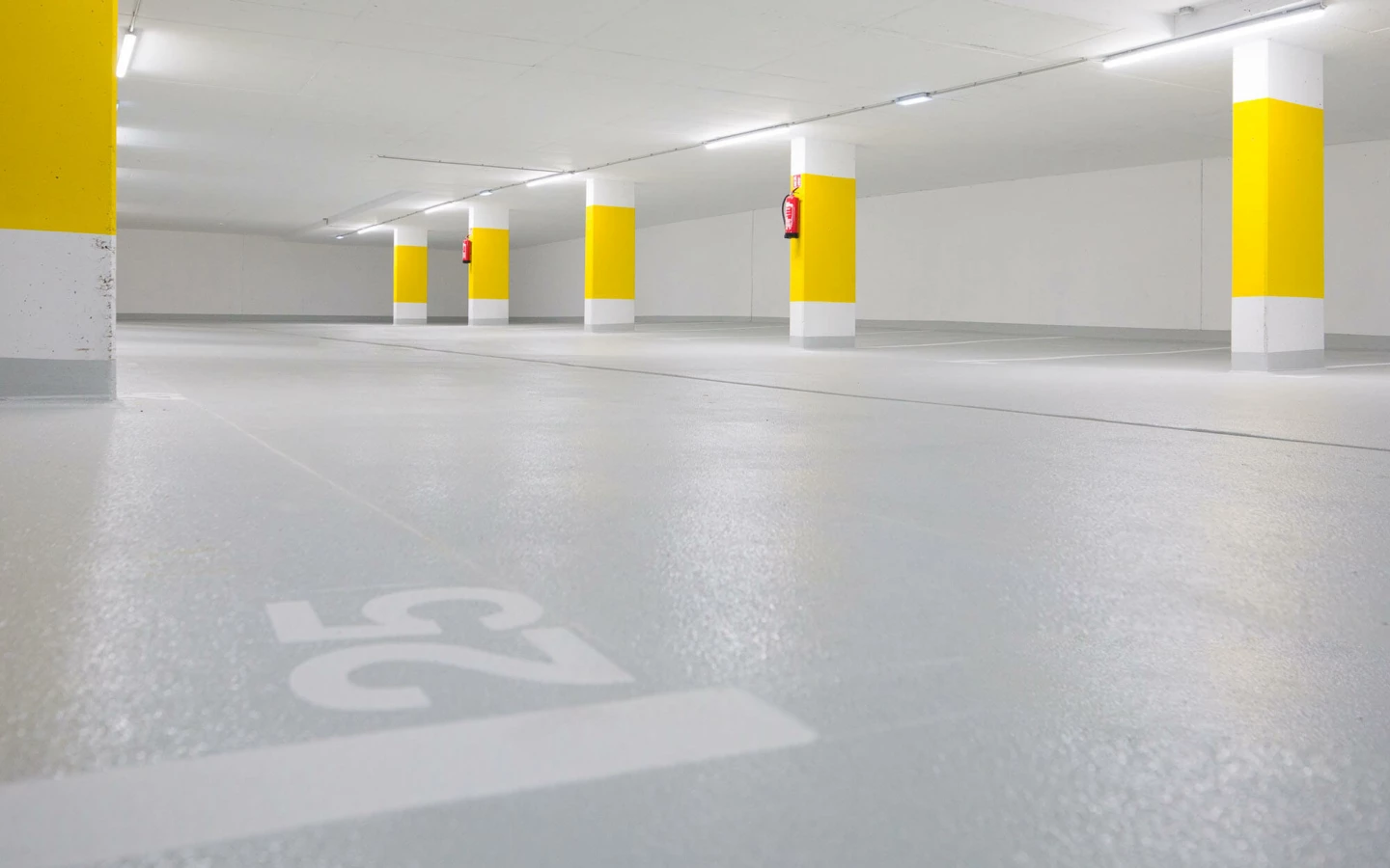 Leere, beleuchtete Tiefgarage mit vielen gelb und weiß gestrichenen Säulen und grauen Stellplätzen, Nummer 25 auf dem Boden (Widum, Wohnanlage, Lustenau).