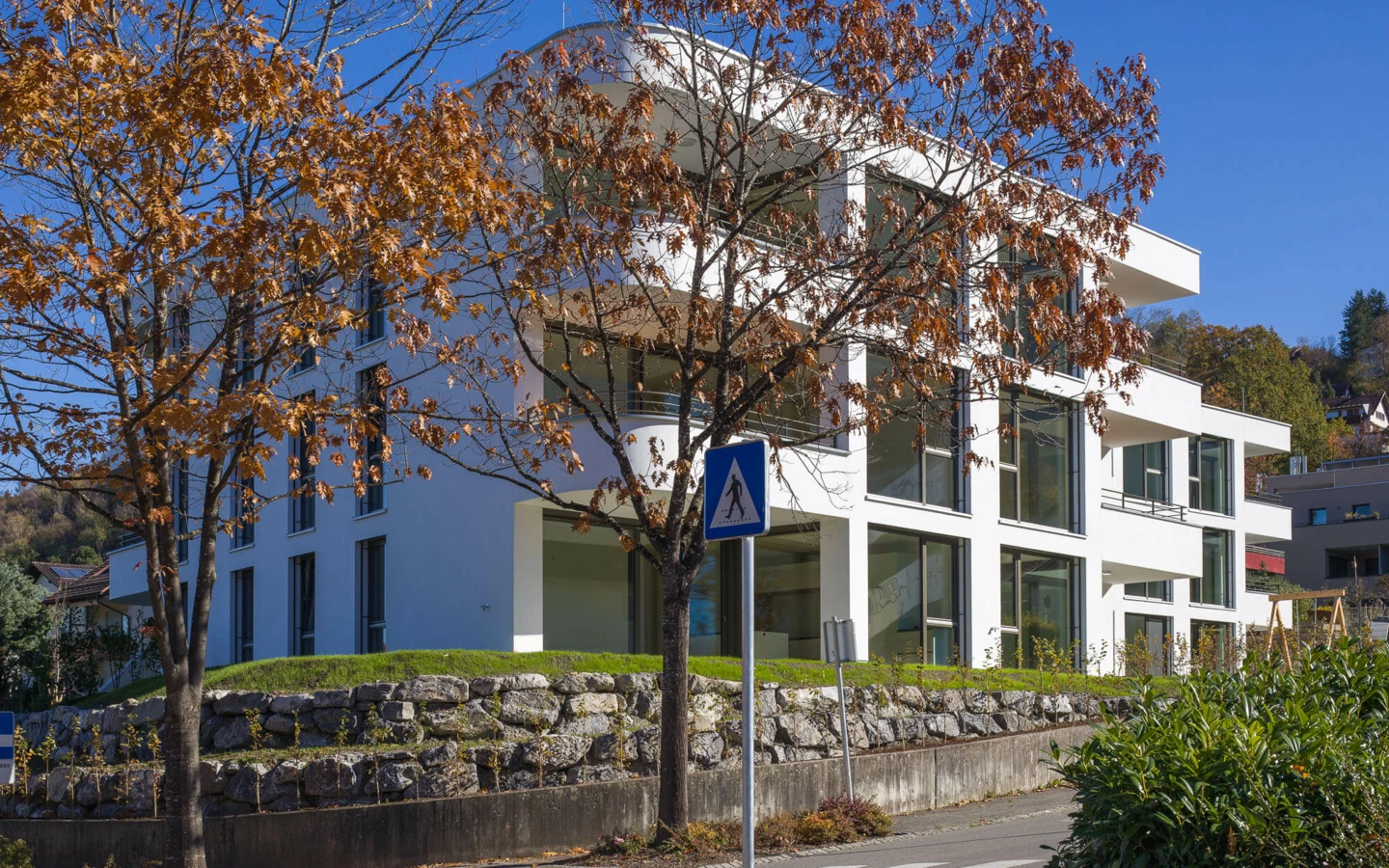 Moderne weiße Wohnanlage an der Wolf-Huber-Straße in Feldkirch mit großen Fenstern und Balkonen, umgeben von Bäumen und einer Steinmauer.