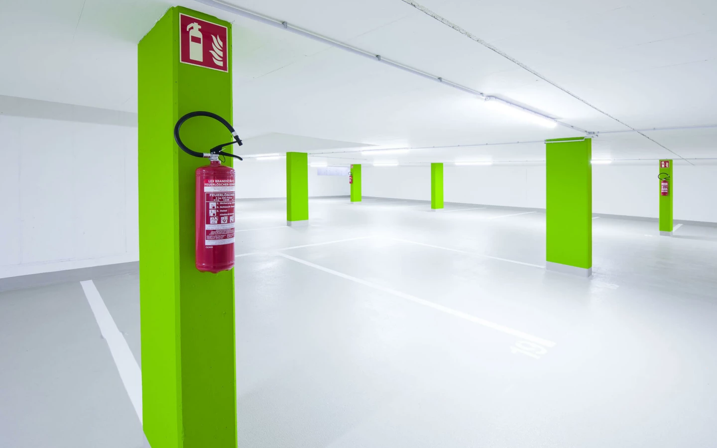Leere, beleuchtete Tiefgarage mit hellgrünen Säulen, Feuerlöschern und weißen Parkplatzmarkierungen (Wolf-Huber-Straße, Wohnanlage, Feldkirch)