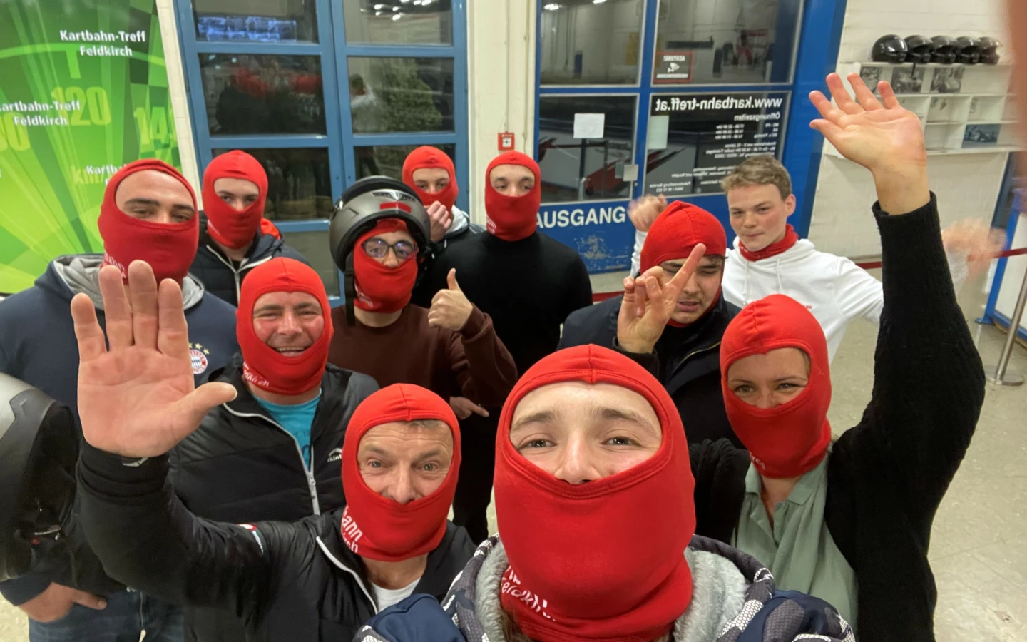 Eine Gruppe fröhlicher Personen trägt rote Sturmhauben und posiert für ein Selfie vor einem Schild mit der Aufschrift "Kartbahn-Treff Feldkirch" und "Ausgang" (Lehrlingsabend).