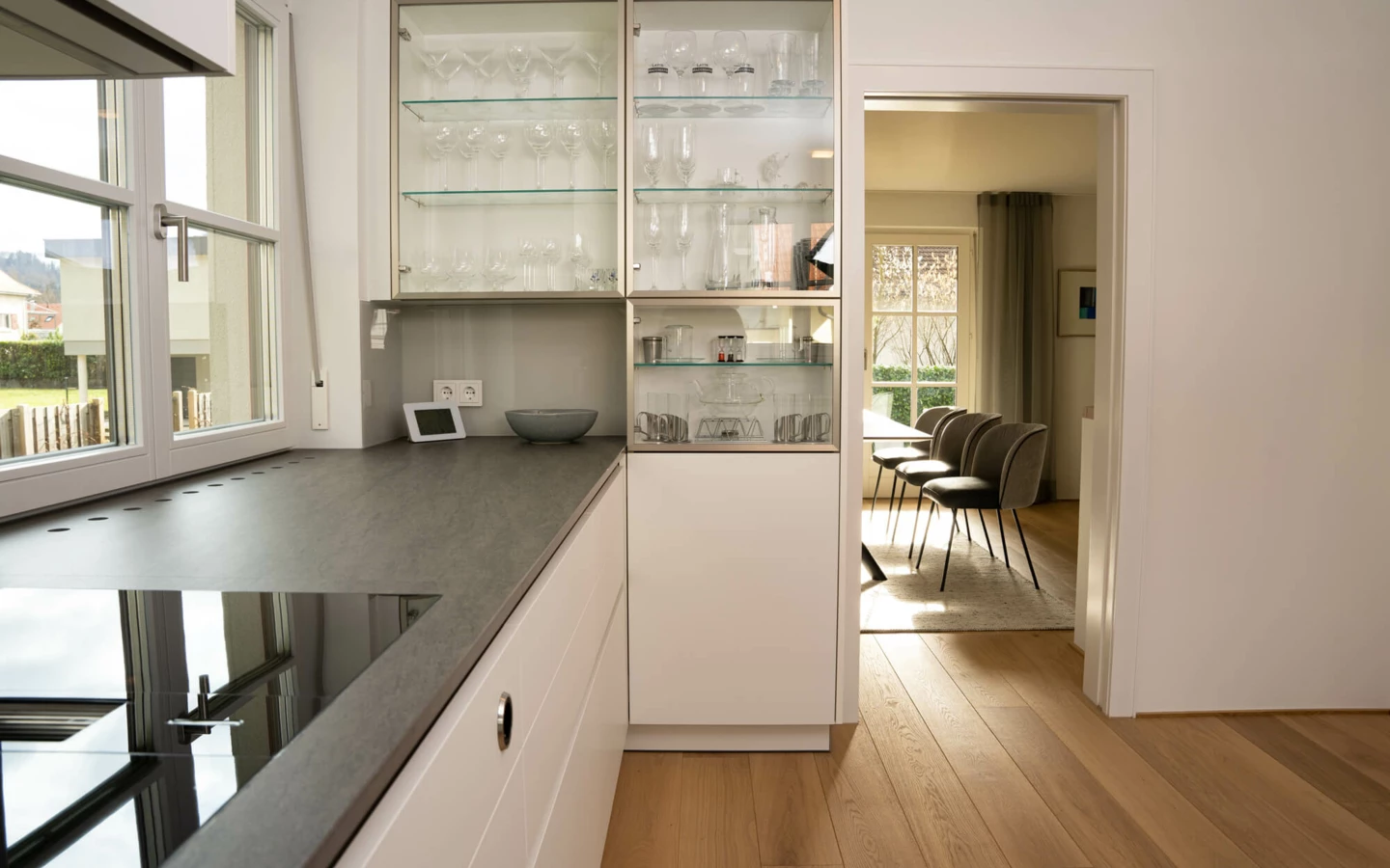 Moderne weiße Küche mit dunkler Arbeitsplatte und Induktionskochfeld, inklusive Glasvitrinenschrank und Blick ins Esszimmer mit Holzboden (Generalsanierung, Bohle).