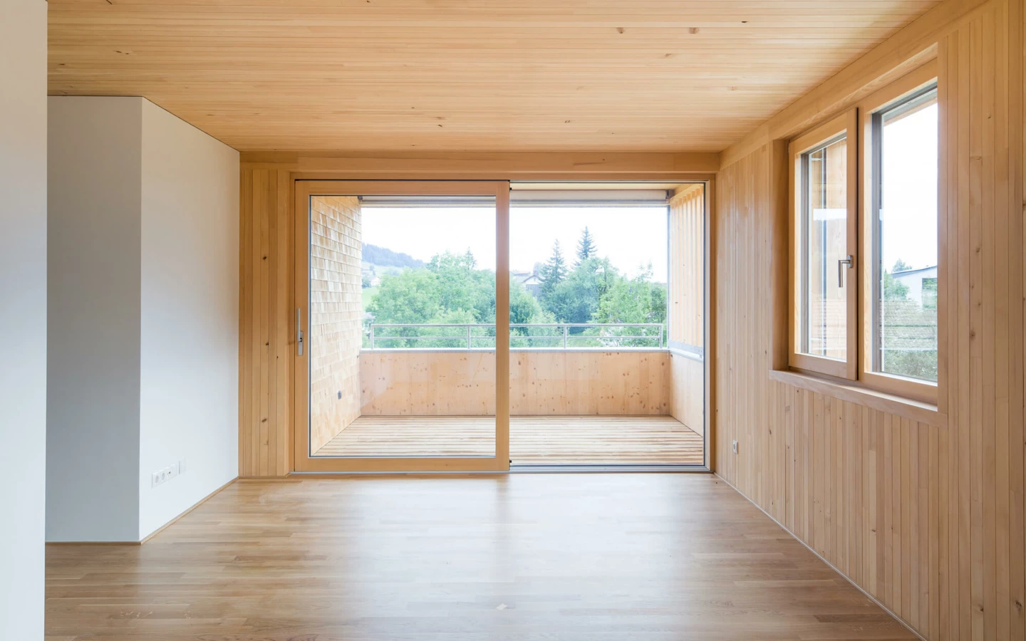 Leerer Raum mit Holzboden und Holzdecke, grosser Schiebetür zum Balkon und Fenster mit Blick ins Grüne (Wohnanlage, Alberschwende)
