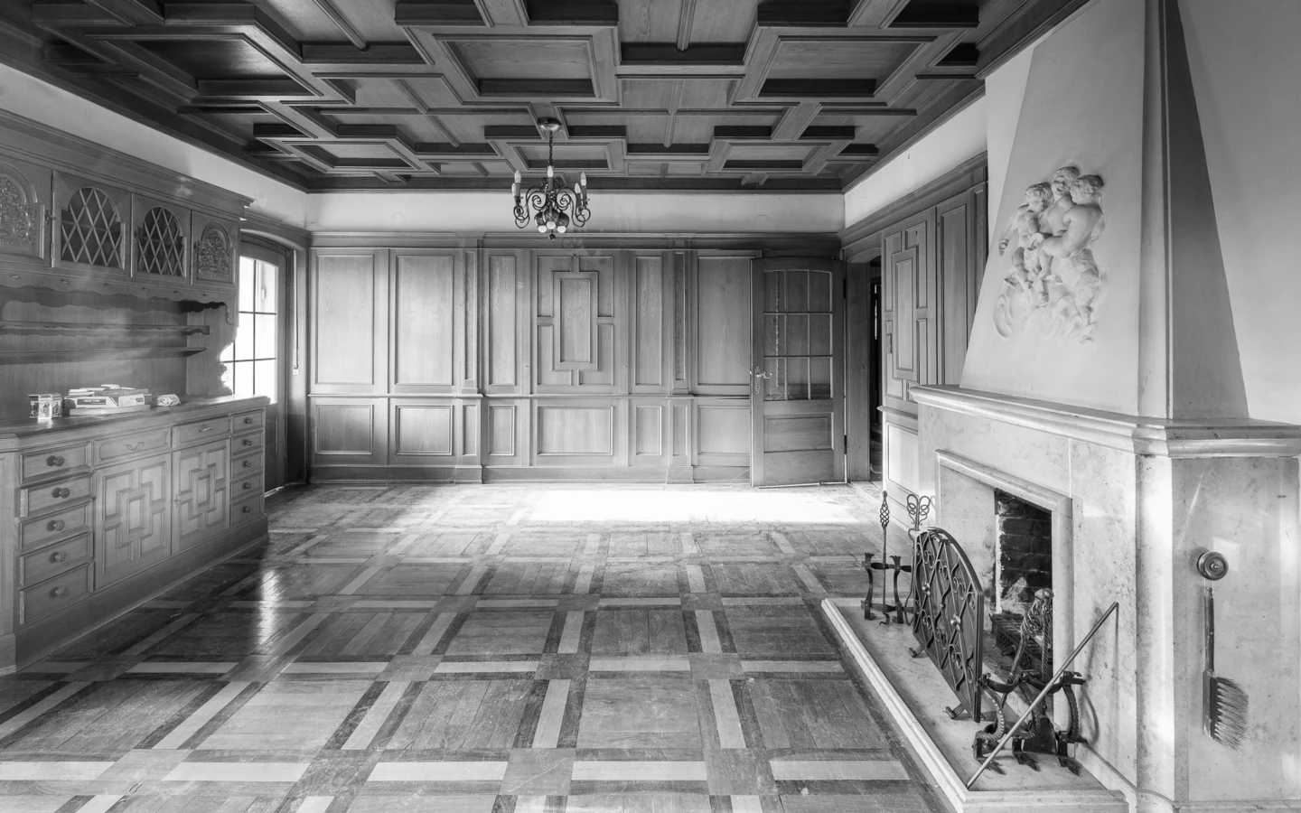 Schwarz-Weiß-Ansicht eines leeren Raumes mit Holzpaneelen, Kassettendecke, Kamin und Parkettboden, dazu ein Kronleuchter (Villa, Reichsstr).