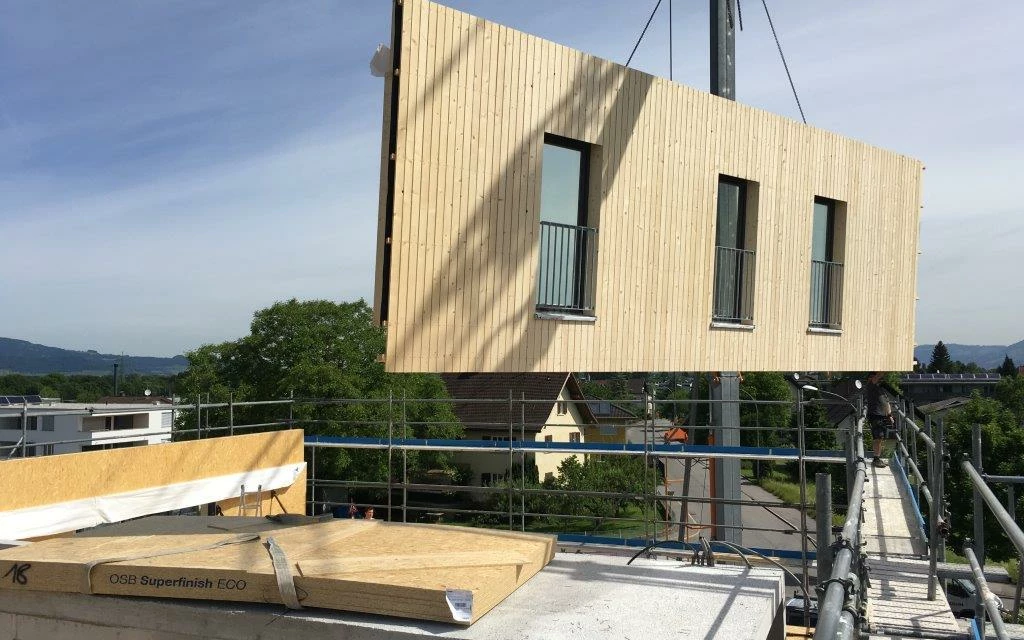 Ein Kran hebt ein vorgefertigtes Holzbaumodul mit drei Fenstern und Balkonen auf eine Baustelle (Architekturexkursionen, Holzbauprojekte)