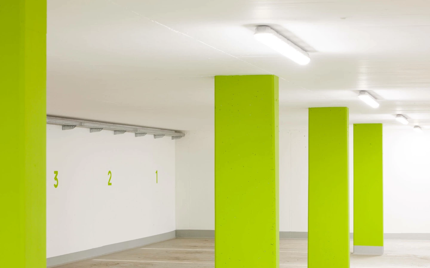 Leere Tiefgarage mit leuchtend grünen Säulen, weiß gestrichenen Wänden und LED-Beleuchtung an der Decke (Feldgasse, Wohnanlage, Dornbirn).