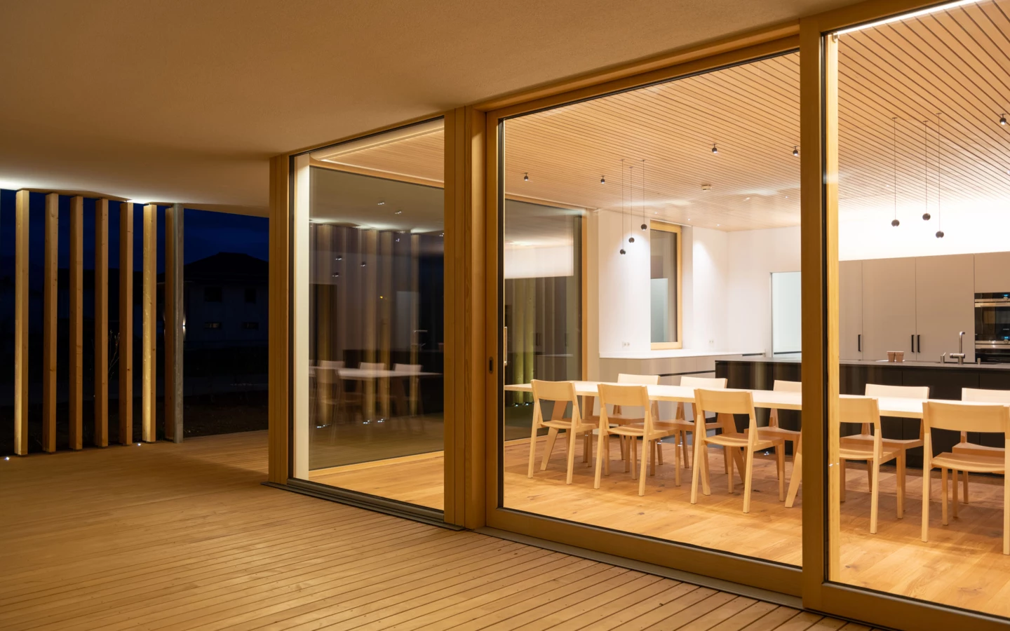 Modernes Esszimmer und Küche mit Holzboden und Glasschiebetüren, die zu einer beleuchteten Holzterrasse führen (Wolfurt, Dieter).