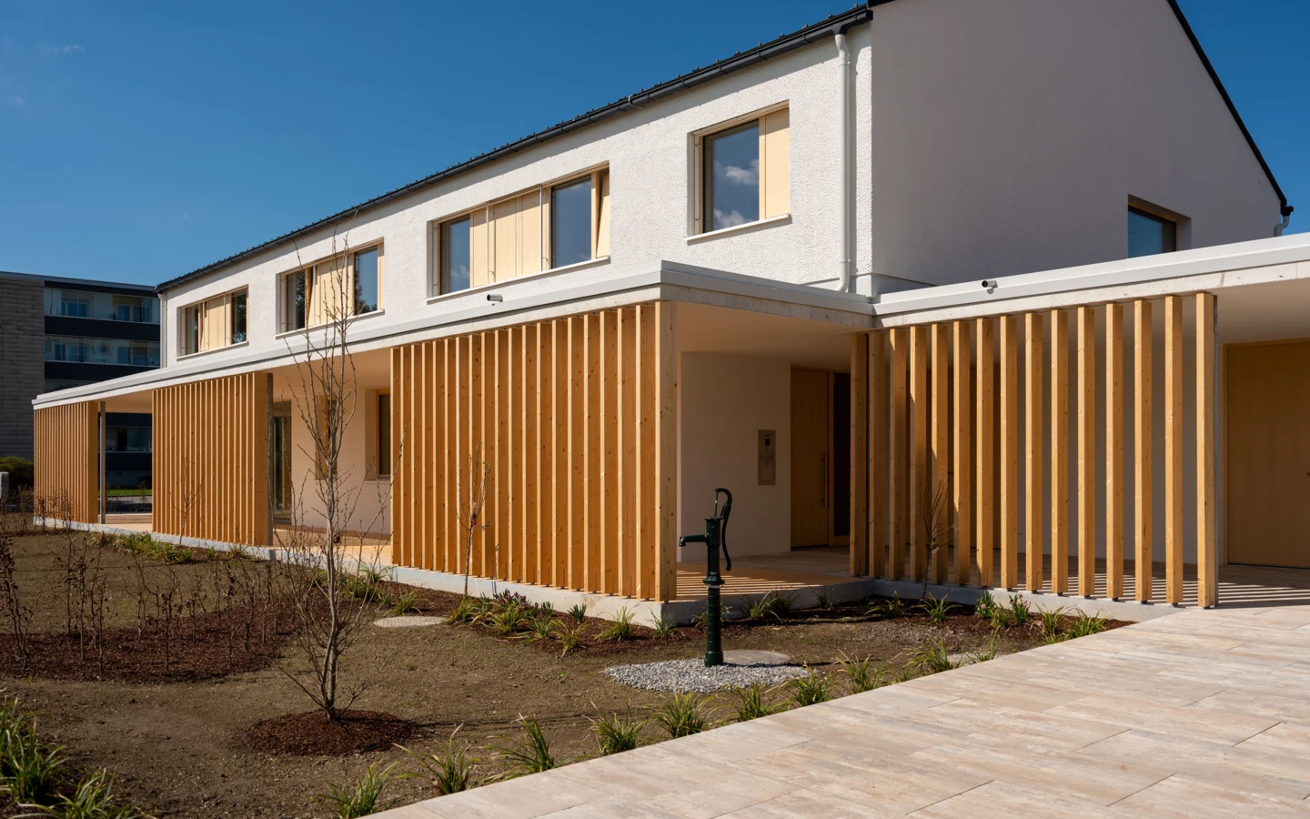 Modernes Wohnhaus mit weißer Fassade und Holzlamellen, im Vordergrund ein Garten mit Gießkanne und gepflastertem Weg (Wolfurt, Dieter).