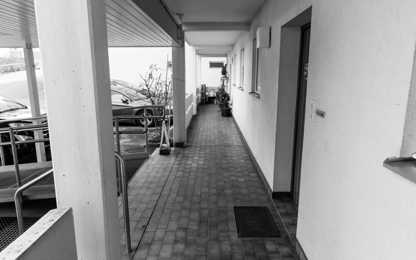 Eine schwarz-weiße Aufnahme eines langen, schmalen Flurs mit Fliesenboden, einer Reihe von Türen auf der rechten Seite, und Autos, die draußen auf der linken Seite geparkt sind (Römerstraße).