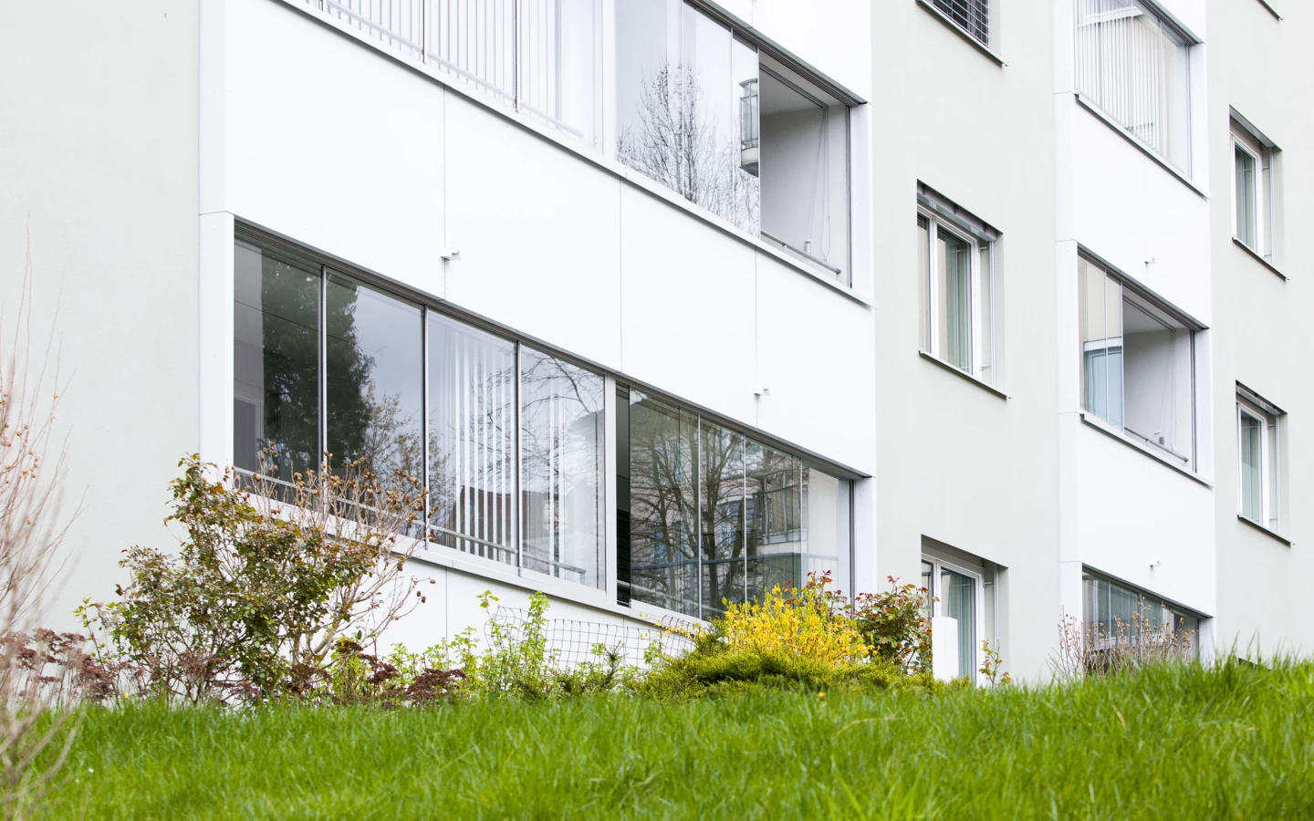 Umfassender Blick auf die weiße Fassade eines modernen Mehrfamilienhauses mit zahlreichen Balkonen und Fenstern, eingebettet in grüne Pflanzen und Gras (Römerstraße).