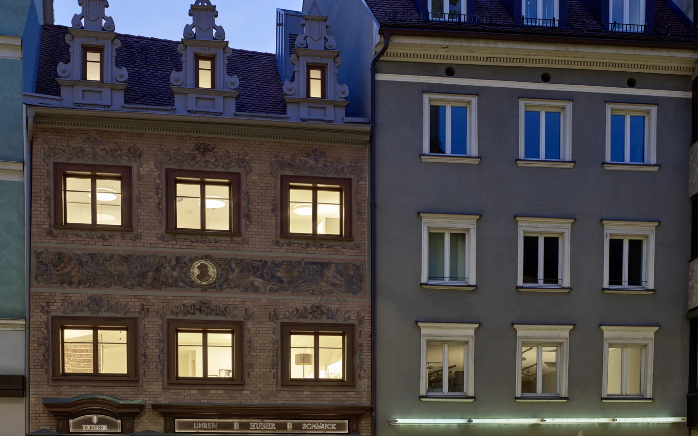 Zwei beleuchtete mehrstöckige Gebäude in der Rathausstraße 7 während der Dämmerung, eines mit 'Uhren Huber' Beschriftung und Schaufenstern.