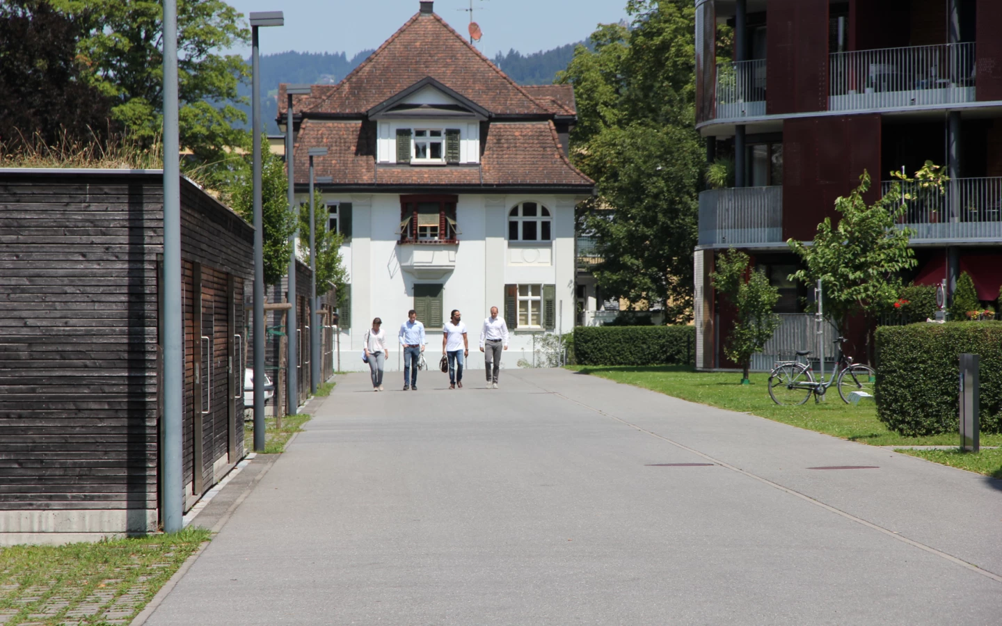 Vier Personen gehen eine Straße entlang, mit einem traditionellen weißen Haus und modernen Gebäuden im Hintergrund.